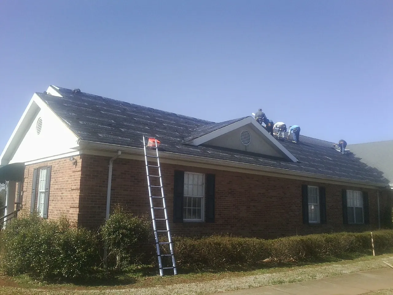 Crew tearing shingles off a brick ranch roof with a ladder set — Metal Roof Repair work by our crew in Halls