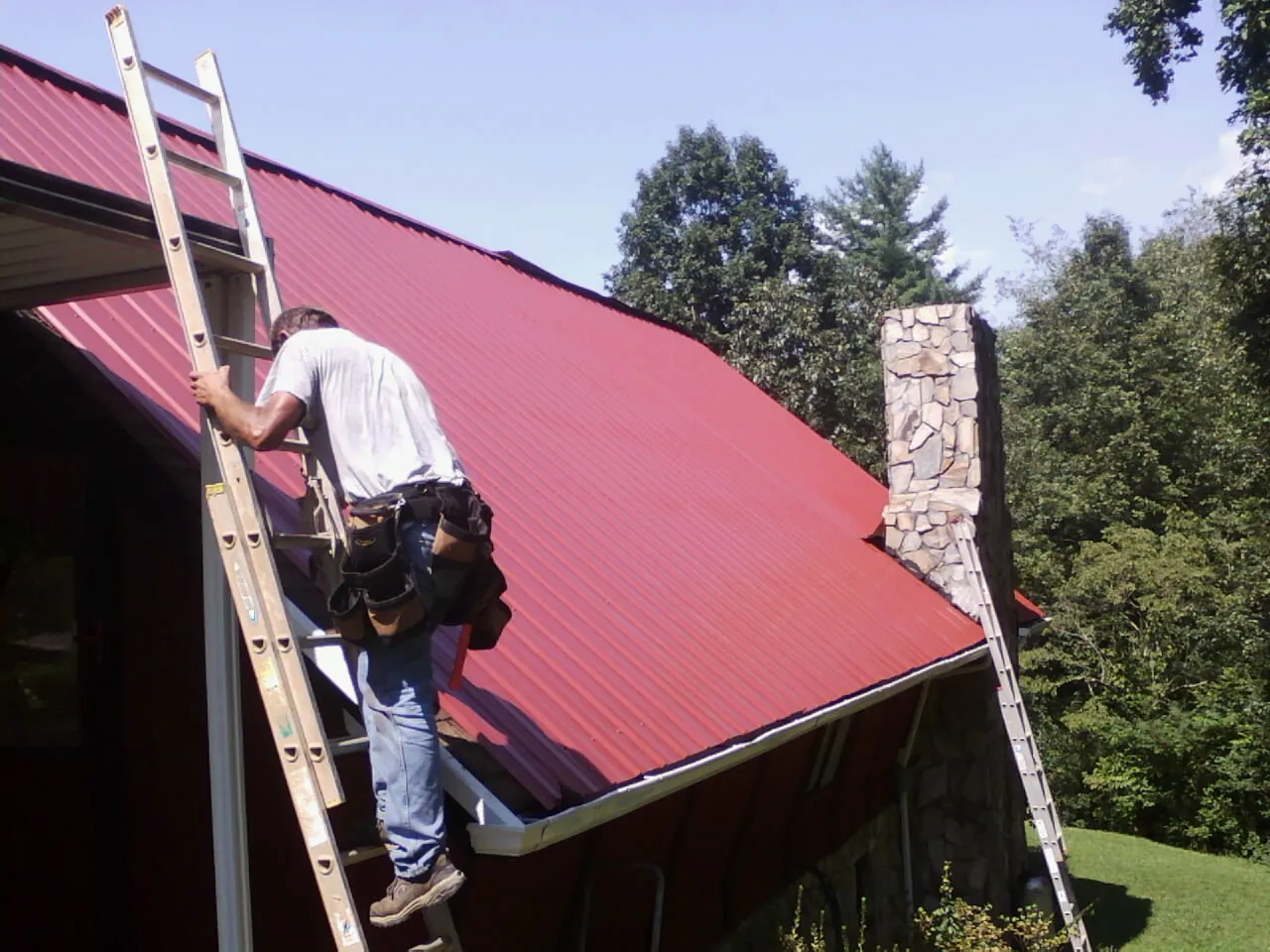 Roofer on a ladder beside a red metal roof and stone chimney — Soffit & Fascia Repair by Roofing Brothers in Halls